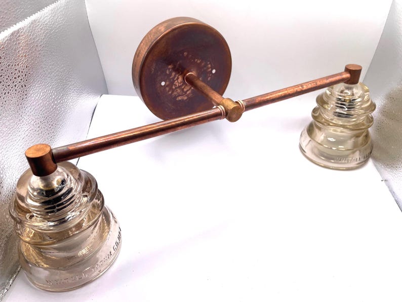 May include: A wall-mounted light fixture featuring a horizontal copper-toned metal bar attached to a circular backplate. Two clear, tiered glass insulators are secured to the ends of the bar, serving as the light shades for this industrial-style lamp.