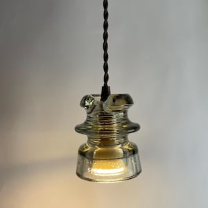 Könnte beinhalten: Eine gläserne Pendelleuchte mit einem braunen, gedrehten Kabel. Das Glas ist hell bernsteinfarben und trägt den Text "WHITTALL  125V  515" auf der Unterseite.