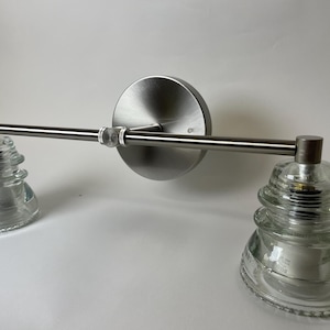 May include: A wall-mounted light fixture featuring a brushed nickel horizontal bar attached to a circular backplate. Two clear, tiered glass insulators serve as shades, each secured to the ends of the metal arm with visible hardware.