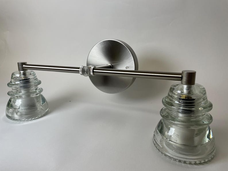 May include: A wall-mounted light fixture featuring a brushed nickel horizontal bar attached to a circular backplate. Two clear, tiered glass insulators serve as shades, each secured to the ends of the metal arm with visible hardware.