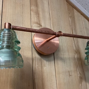 May include: A wall-mounted light fixture featuring a horizontal copper pipe arm attached to a circular copper base. Two light sockets are fitted with translucent, pale green, tiered glass electrical insulators, each with a wide, ridged base.