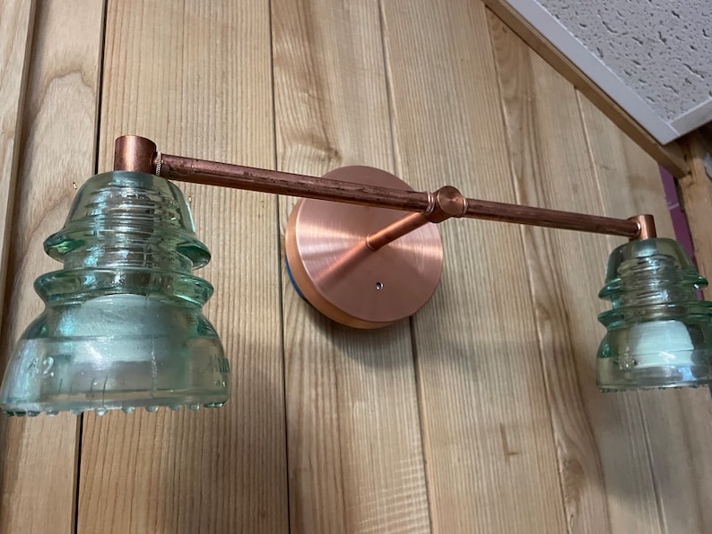 May include: A wall-mounted light fixture featuring a horizontal copper pipe arm attached to a circular copper base. Two light sockets are fitted with translucent, pale green, tiered glass electrical insulators, each with a wide, ridged base.