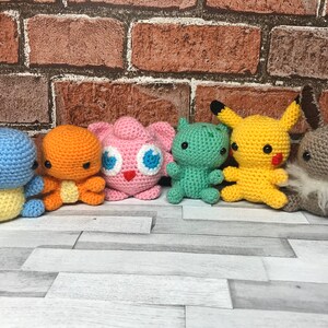 May include: A collection of six crocheted Pokémon figures. The figures include Squirtle, Charmander, Jigglypuff, a green creature, Pikachu, and Eevee. Each figure is brightly colored and has distinct features, with black eyes and embroidered details.
