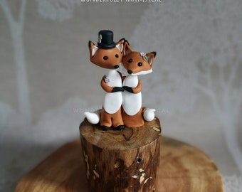 Pode incluir: Um topo de bolo com dois raposas, uma delas usando um chapéu de copa, em pé sobre um tronco de madeira. As raposas são feitas de fondant marrom, branco e laranja. O texto "CAKE TOPPERS By WONDERFULLY WHIMSICAL" está acima das raposas.