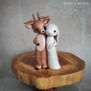 May include: A wooden cake topper featuring a brown deer with antlers and a white rabbit. The deer is holding the rabbit close. The topper is on a wooden base. "Cake Toppers by Wonderfully Whimsical" is written on the background.