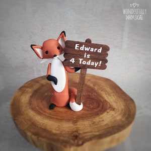 Pode incluir: Uma figura de raposa vermelha segurando uma placa de madeira que diz "Edward is 4 Today!" A figura está em um suporte de bolo de madeira.