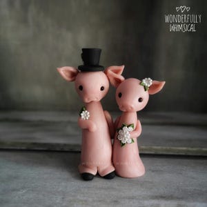 May include: Two pink pig figurines standing side by side. The left pig wears a black top hat and has a small white flower on its shoulder. The right pig has a white flower on its ear and holds a bouquet of white flowers. Text reads WONDERFULLY WHIMSICAL.