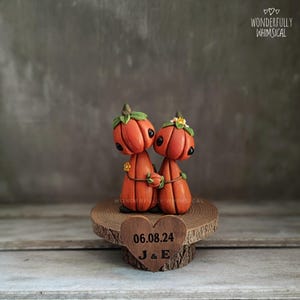 May include: A pair of orange pumpkin-shaped figurines with black eyes and green leafy tops, holding hands, atop a wooden base. The base has a heart-shaped plaque with the date 06.08.24 and initials J & E. The words "Wonderfully Whimsical" are in the top right corner.