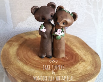 Pode incluir: Um topo de bolo de casamento com um casal de ursos marrons com flores cor-de-rosa. A ursa noiva está usando uma coroa de flores e o urso noivo está segurando um buquê. O topo de bolo está em uma base de madeira com o texto "Cake Toppers by Wonderfully Whimsical".