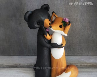 Pode incluir: Um topo de bolo de um urso preto e uma raposa. O urso está abraçando a raposa, que está usando uma coroa de flores. O topo de bolo é feito de argila polimérica e é pintado à mão. Diz "CAKE TOPPERS by WONDERFULLY WHIMSICAL" no topo.