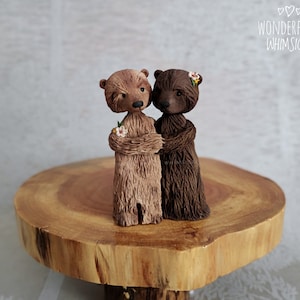 May include: Two brown bear figurines, one light brown and one dark brown, stand close together. Each bear has a small flower bouquet. The figurines are on a wooden slice, with the text "Wonderfully Whimsical" in the upper right corner.