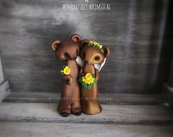 Pode incluir: Um topo de bolo de casamento com um casal de ursos marrons. A ursa noiva está usando um vestido branco com uma coroa de flores amarelas e um buquê de flores amarelas. O urso noivo está usando um terno marrom e segurando uma única flor amarela.