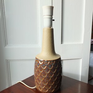 May include: A ceramic table lamp with a beige neck and a textured, brown and blue patterned base. The lamp has a white bulb socket and a power cord. The lamp is on a dark wooden table.