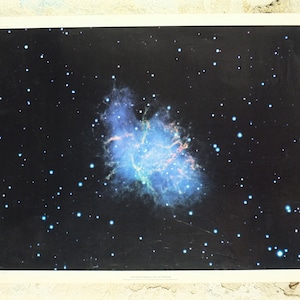 May include: A poster showing the Crab Nebula, a celestial object, set against a black backdrop speckled with white stars. The nebula is a swirling mass of blue, green, and orange colours.
