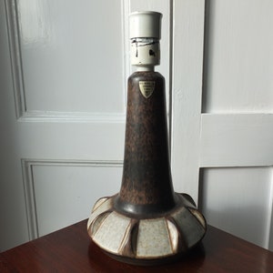 May include: A vintage table lamp with a brown speckled body and a white top. The base features a decorative pattern with alternating sections of brown and off-white. The lamp is placed on a dark wooden surface.