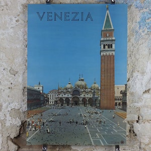 May include: A vintage travel poster featuring the city of Venice, Italy. The poster displays the word "VENEZIA" in blue against a clear sky. The image showcases St. Mark's Campanile and the Basilica, with people in the square.