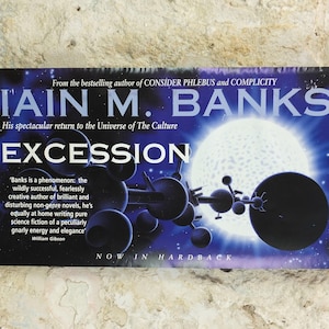 Puede incluir: Un cartel promocional de la novela "Excession" de Iain M. Banks. El título está en letras blancas grandes, con el nombre del autor encima. La portada presenta una escena espacial azul oscuro y púrpura con una gran esfera blanca y una estructura compleja de color oscuro.