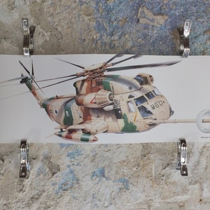 May include: A detailed illustration of a military helicopter in camouflage colours, featuring tan, green, and brown. The helicopter, marked with "960", is secured to a textured wall with metal clips. The image shows the helicopter's rotor.