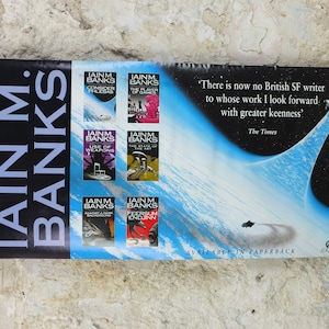 Puede incluir: Póster con portadas de libros de Iain M. Banks, incluyendo "Consider Phlebas" y "The Player of Games". El póster tiene un fondo azul y negro con temática espacial y el texto "Available in Paperback".