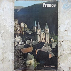 May include: A vintage travel poster for France, featuring a picturesque view of a village with a church and tall spires. The word "France" is printed at the top. The poster is displayed against a distressed, textured wall.
