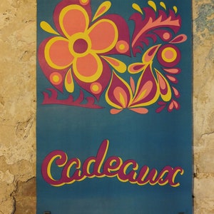 Pu&ograve; includere: Un poster turchese con un design floreale nei toni del rosa, arancione e giallo. La parola "Cadeaux" &egrave; scritta in corsivo con un contorno giallo e un riempimento rosa. Il poster &egrave; tenuto da clip metalliche.