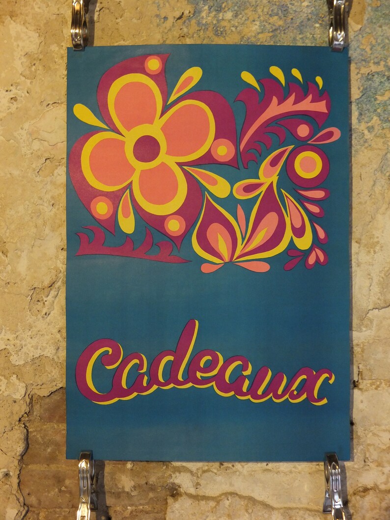 Pu&ograve; includere: Un poster turchese con un design floreale nei toni del rosa, arancione e giallo. La parola "Cadeaux" &egrave; scritta in corsivo con un contorno giallo e un riempimento rosa. Il poster &egrave; tenuto da clip metalliche.