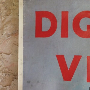 Puede incluir: Primer plano de un p&oacute;ster con la palabra "DIG" en letras rojas grandes, y la letra "V" en rojo. El p&oacute;ster es de color azul claro y est&aacute; sujeto por una pinza plateada. El fondo es una pared de ladrillo marr&oacute;n claro texturizada.