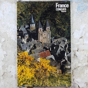 May include: A vintage travel poster featuring a scenic view of Conques, France. The poster displays a medieval town with a church and dark rooftops, surrounded by lush greenery and yellow flowers. The text "France CONQUES Aveyron" is at the top.