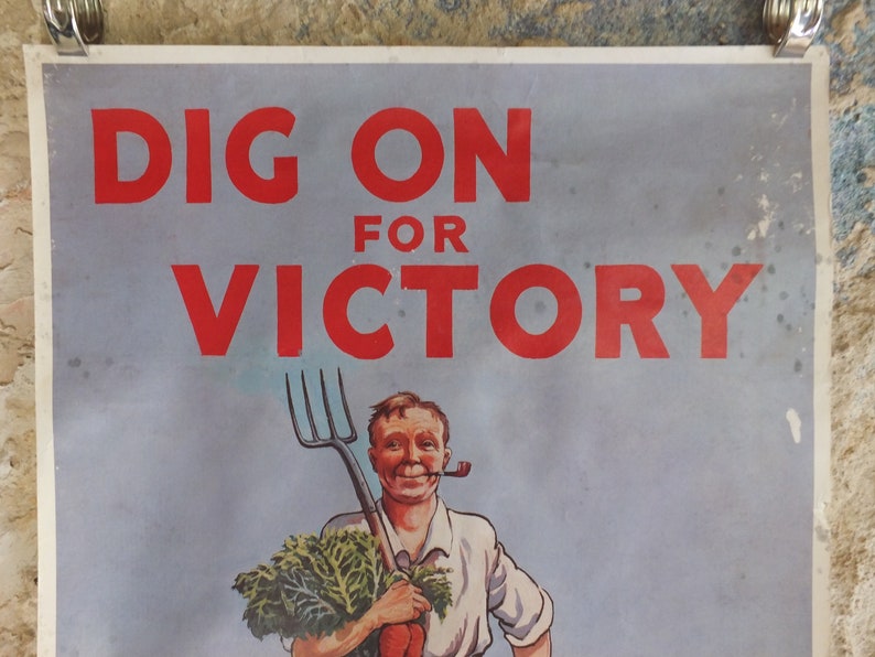 Puede incluir: Cartel vintage con el texto "DIG ON FOR VICTORY" en rojo. La imagen muestra a un hombre sonriente con una pipa, sosteniendo un horca y verduras. El fondo es azul claro, con pinzas met&aacute;licas.