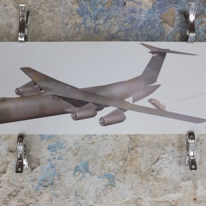 May include: A print of a Lockheed C-141 Starlifter cargo plane, in shades of gray and olive green, is held by metal clips against a textured, off-white wall. The aircraft is depicted in profile.