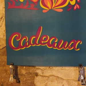 Pu&ograve; includere: Un cartello turchese con la parola "Cadeaux" in un carattere corsivo a strati, con un contorno giallo e un riempimento magenta. Il cartello &egrave; decorato con ritagli floreali colorati e tenuto da clip metalliche contro un muro di mattoni strutturato.