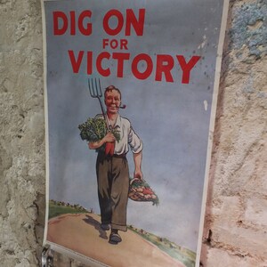 Puede incluir: Cartel vintage con el texto "DIG ON FOR VICTORY" en rojo. La imagen muestra a un hombre sonriente con una pipa, sosteniendo una horca y una cesta de verduras. El cartel est&aacute; sujeto con pinzas met&aacute;licas contra una pared texturizada.