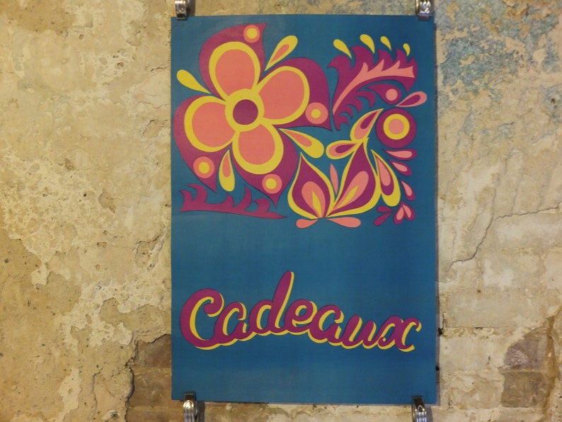 Pu&ograve; includere: Un poster turchese con un design floreale rosa, giallo e viola. La parola "Cadeaux" &egrave; scritta in un font stilizzato in basso. Il poster &egrave; montato su un muro strutturato marrone chiaro.