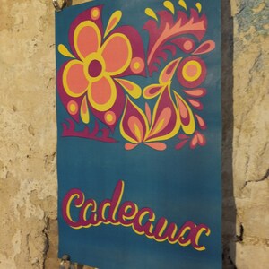 Pu&ograve; includere: Un poster turchese con un design floreale rosa, giallo e arancione. La parola "Cadeaux" &egrave; scritta in corsivo rosa con un contorno giallo. Il poster &egrave; tenuto da clip metalliche.