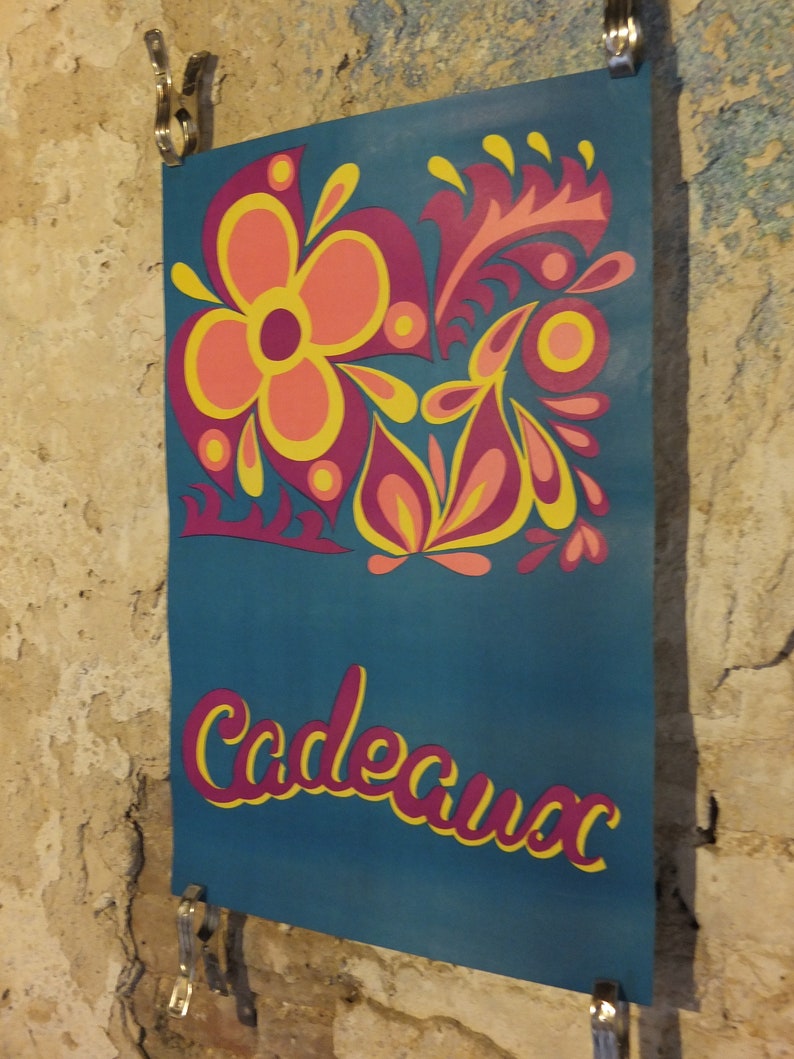 Pu&ograve; includere: Un poster turchese con un design floreale rosa, giallo e arancione. La parola "Cadeaux" &egrave; scritta in corsivo rosa con un contorno giallo. Il poster &egrave; tenuto da clip metalliche.