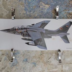 Può includere: Una stampa di un jet militare grigio e nero, con insegne tedesche e il numero 46+13. L'immagine è tenuta da clip argentate contro un muro strutturato di colore chiaro. Il jet ha un design dettagliato.