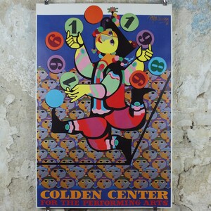 May include: A colorful poster featuring a stylized figure juggling numbered circles. The figure is depicted in bright colors against a blue background, with a crowd of faces at the bottom. The poster promotes the Golden Center for the Performing Arts.