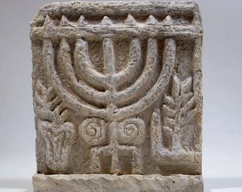 May include: A white stone relief sculpture of a menorah with a decorative border. The menorah is surrounded by stylized floral designs.