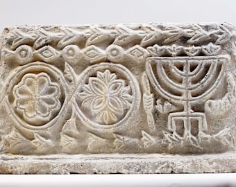 May include: A rectangular, off-white stone carving featuring floral and geometric designs. The carving includes a menorah, floral patterns, and leaf motifs, showcasing a detailed, textured surface. The overall design suggests a historical or religious theme.