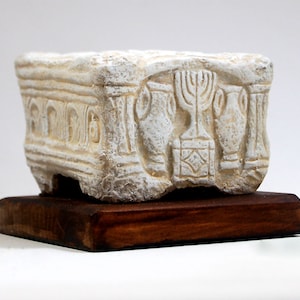 May include: A white stone sculpture of a menorah, two cups, and a small building with a rounded roof. The sculpture is on a wooden base.