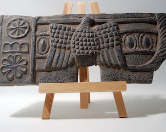 May include: A dark gray stone relief sculpture of a winged creature with a circular design on the left side. The sculpture is displayed on a wooden easel.