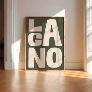 Könnte beinhalten: Ein gerahmter Kunstdruck mit dem Wort "LAGANO" in großen, cremefarbenen Blockbuchstaben auf einem dunkelgrünen Hintergrund. Der Rahmen ist dünn und goldfarben. Das Kunstwerk steht auf einem Holzboden und lehnt an einer weißen Wand.
