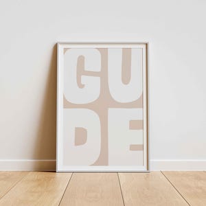 以下が含まれることがあります： 明るい茶色の背景に、大きく丸みを帯びた白い文字で「GUDE」と書かれた額入りのアートプリント。 白いフレームに入ったアートワークは、木製の床に白い壁に立てかけられています。