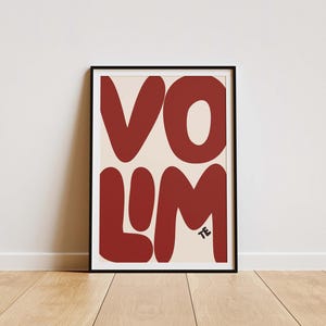Könnte beinhalten: Ein gerahmter Kunstdruck mit dem Wort "VOLUM" in großen, abgerundeten, rostfarbenen Buchstaben auf cremefarbenem Hintergrund. Die Buchstaben sind stilisiert und modern, mit einem kleinen "TE" in der unteren rechten Ecke. Der Rahmen ist schwarz, und der Druck wird auf einem Holzboden präsentiert.