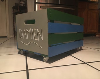 Può includere: Una cassa di legno grigia, verde e blu con ruote. La cassa ha una maniglia sulla parte superiore e il nome "Damien" è scritto sul davanti.