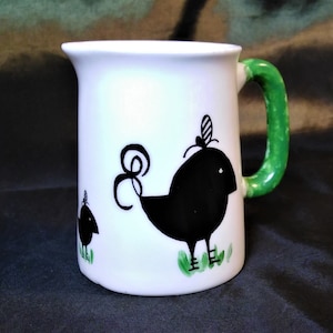 May include: White ceramic pitcher with a green handle. The pitcher features a black and white illustration of two birds. The larger bird has a green leaf tail and a green leaf on its head. The smaller bird is standing on a green leaf.