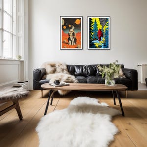 Op de afbeelding: Twee ingelijste illustraties hangen aan een witte muur boven een zwarte leren bank. De illustratie links toont een zwart-witte kat in een ruimtepak die op een rode planeet zit. De illustratie rechts toont een rode figuur die door een bos loopt.