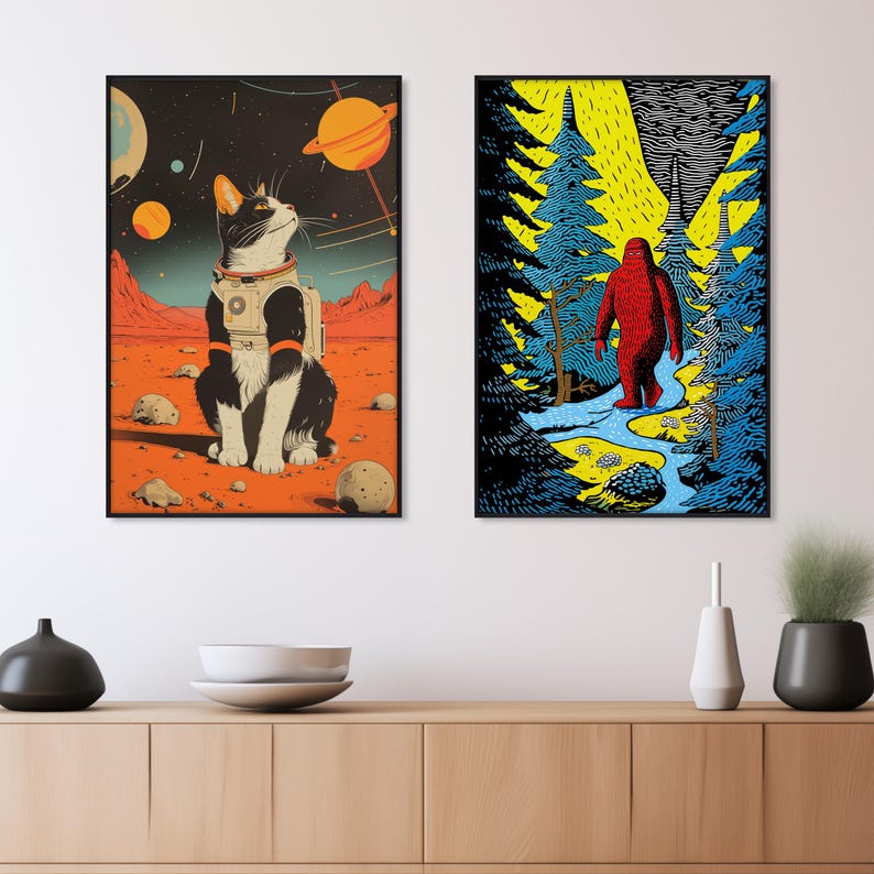 Op de afbeelding: Twee ingelijste illustraties. De eerste toont een zwart-witte kat die een ruimtepak draagt op een rode planeet met een sterrenhemel en planeten op de achtergrond. De tweede toont een rode Bigfoot die door een blauw en groen bos loopt.