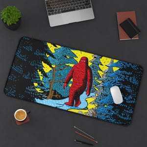 May include: A rectangular black desk mat featuring a red Bigfoot walking through a forest scene with blue and yellow accents. A laptop, mouse, and other office supplies are visible.