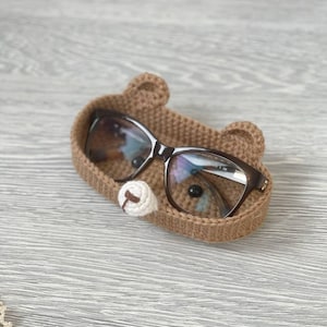 May include: A brown crocheted glasses holder shaped like a bear's face with white felt eyes and nose.